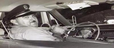 a police officer sitting in the front seat of his car, turning back toward the camera